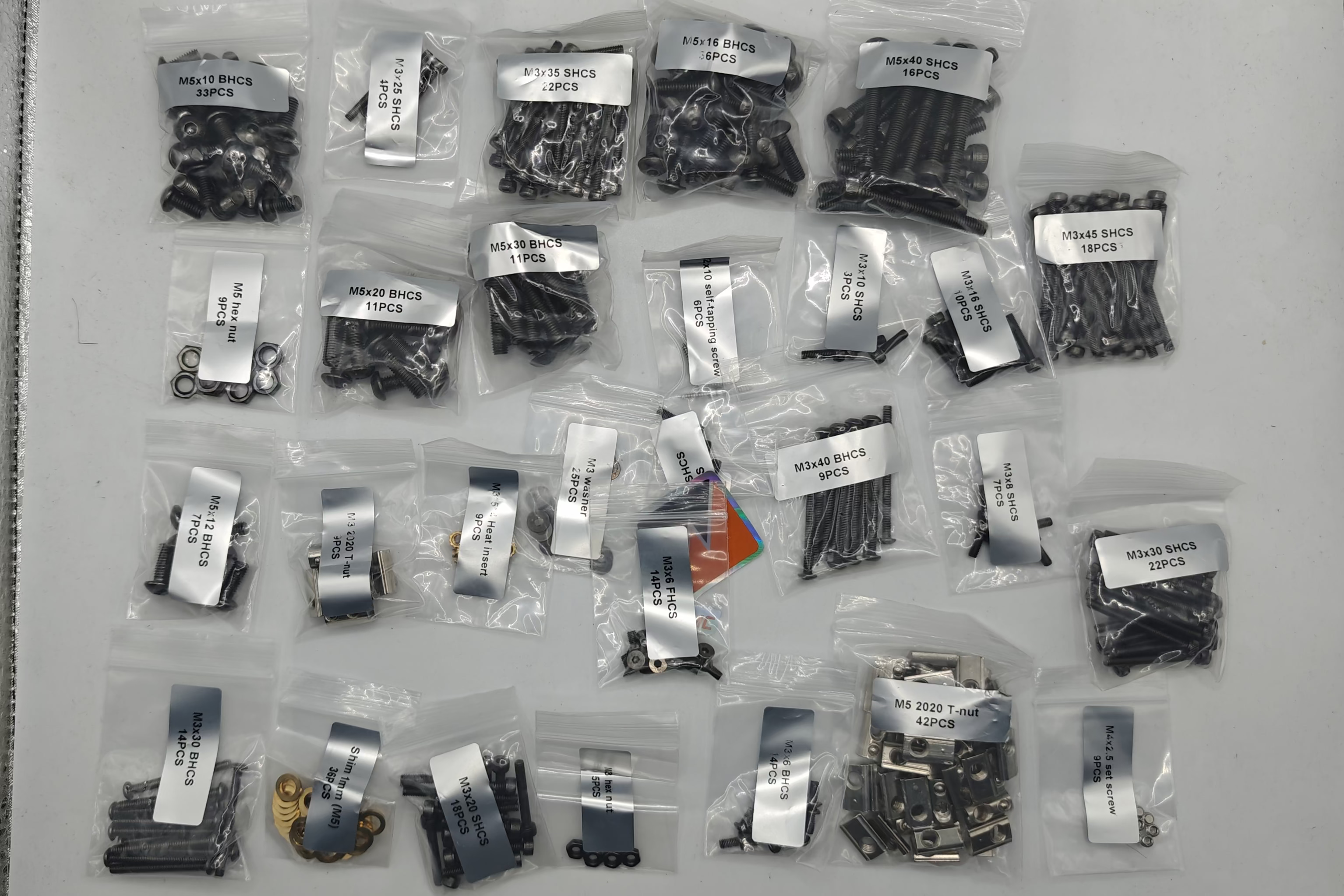 A collection of small clear plastic bags containing various hardware components, including screws, bolts, washers, and clips, each labeled with a white paper tag, arranged on a light gray surface.