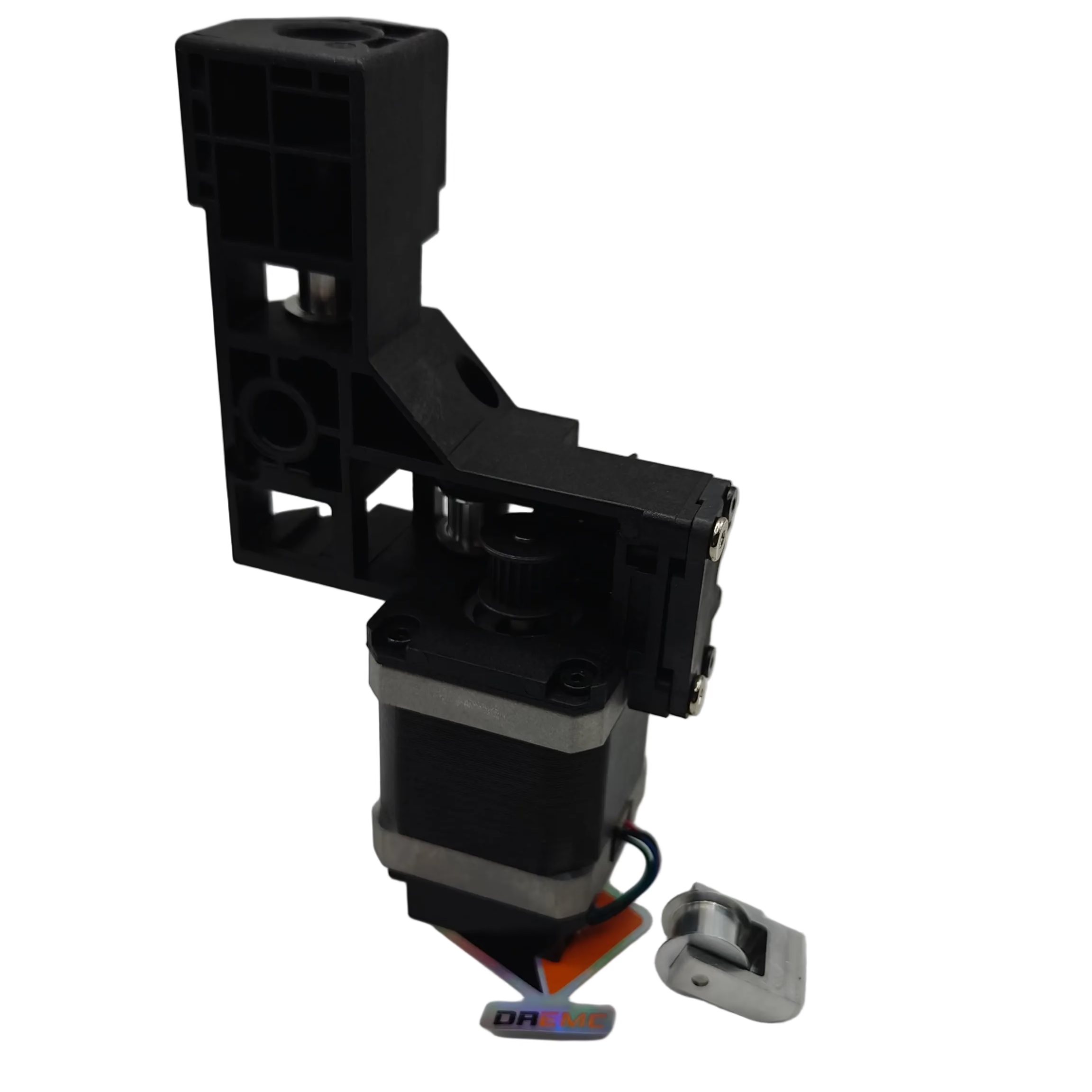 A black plastic mechanical component or bracket assembly with a silver metal base, shown on a plain light gray studio background. A small orange tag and a small white accessory are placed near the base.